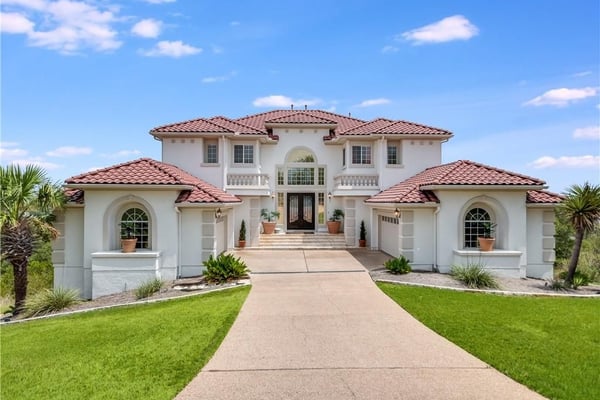 Two-story white stucco mansion with red tile roof, circular arched windows, and manicured lawn with palm trees.
