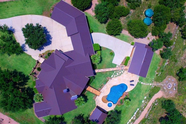 Aerial view of large residential property with purple metal roofs, curved concrete patios, blue swimming pool, and surrounding green lawn and trees