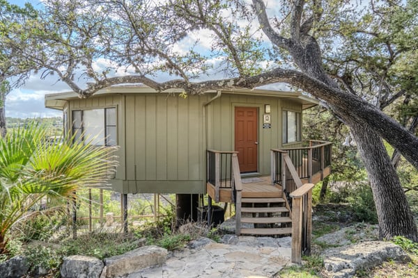 Elevated cabin with tan siding and red door nestled under large oak trees, wooden deck and stairs, scenic hillside views