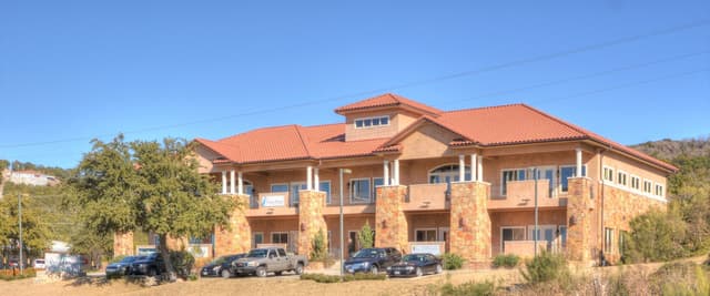Two-story stucco and brick building with orange tile roof, white columns, and multiple windows, parked cars in front, surrounded by desert vegetation and hills.