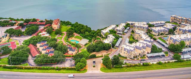 Aerial view of lakeside residential community with red-roofed buildings on left, cream-colored homes on right, green lawns, and calm water beyond.