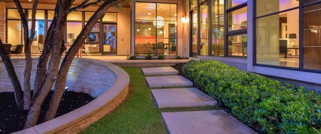 Modern home exterior at dusk with illuminated glass walls, stepping stone pathway through manicured lawn, mature tree, and patio seating visible inside.