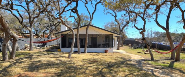 Single-story cottage with tan awning nestled among large oak trees with sparse foliage, red hammocks visible in yard
