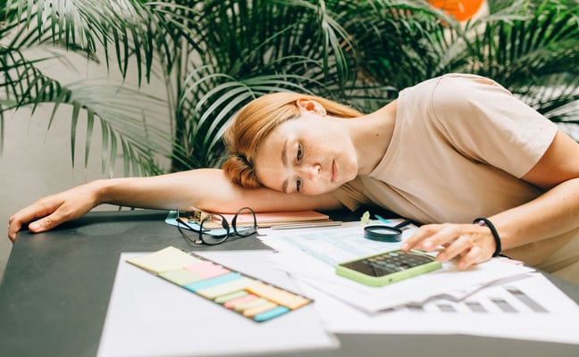 Woman resting head on desk surrounded by papers, calculator, and sticky notes with tropical plants in background