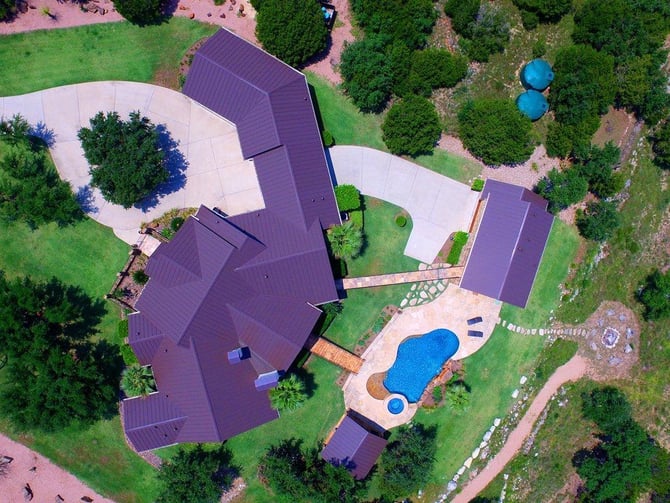 Aerial view of large residential property with purple metal roofs, curved concrete patios, blue swimming pool, and surrounding green lawn and trees