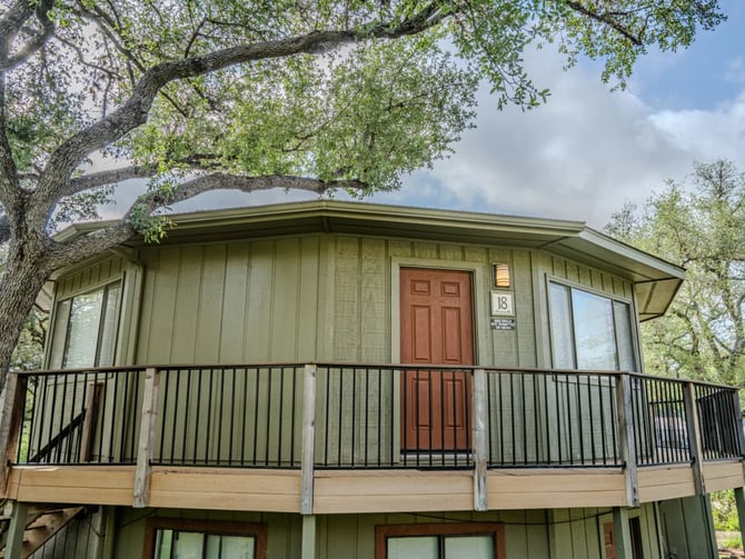 Modern elevated cabin with sage-green siding, red door, and metal railing deck, shaded by large spreading oak tree