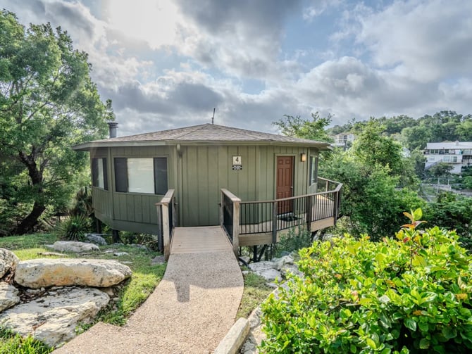 Modern green metal cabin with wraparound deck and stone pathway, surrounded by trees and vegetation