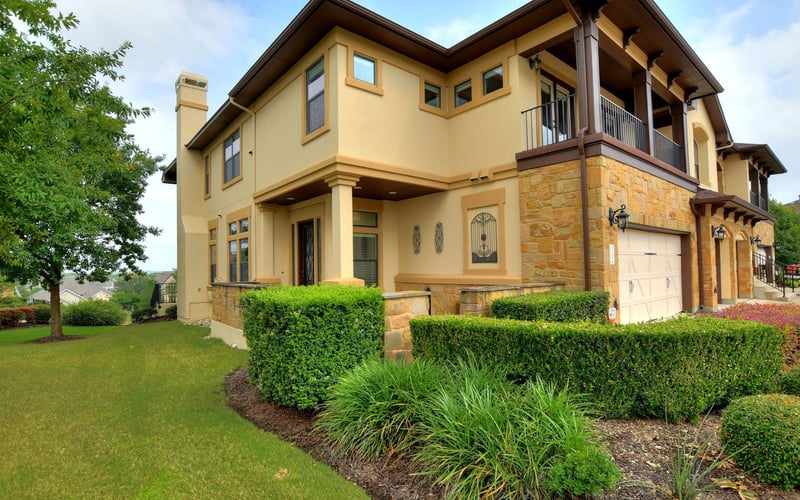 Two-story cream and stone house with dark roof, upper balcony, garage, manicured lawn and hedges