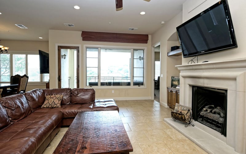 Open-plan living room with brown leather sectional, fireplace, wall-mounted TV, and glass doors overlooking a balcony