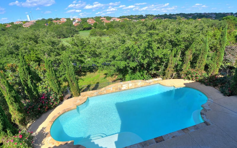 Backyard swimming pool with turquoise water surrounded by cypress trees and manicured landscaping, overlooking suburban neighborhood with red-roofed houses