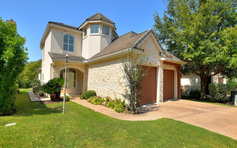Two-story stone and stucco house with tower, red garage doors, manicured lawn, and mature trees under clear blue sky