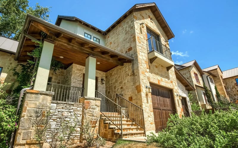 Modern stone house with covered porch, metal railings, and two-story tower under clear blue sky