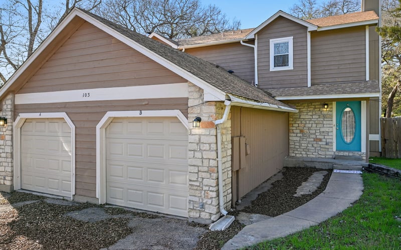 Two-story house with two-car garage, stone and siding exterior, turquoise front door, bare trees in background.