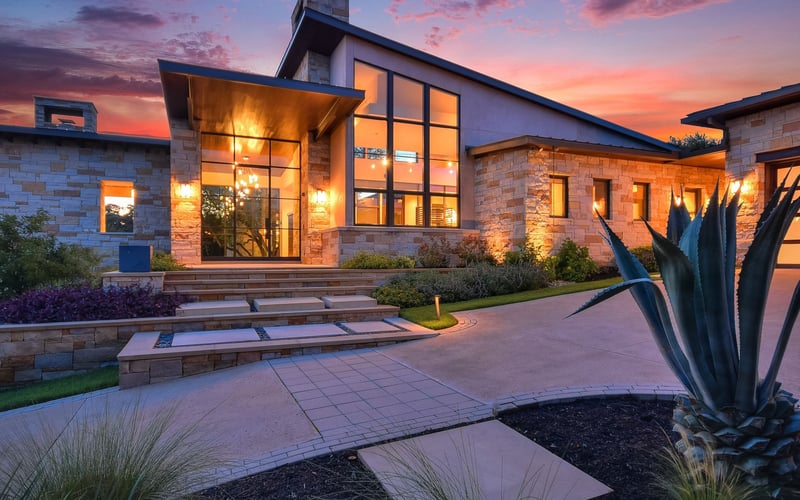 Modern stone house with large windows illuminated at dusk, featuring paved courtyard, desert landscaping, and colorful sunset sky