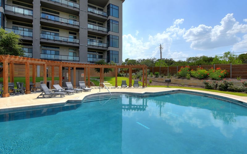 Resort swimming pool with lounge chairs, wooden pergola, and multi-story apartment building in background