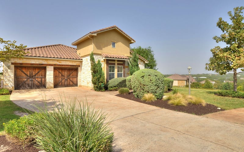 Two-story stone and stucco house with terracotta tile roof, double wooden garage doors, and manicured landscaping on a concrete driveway overlooking rolling