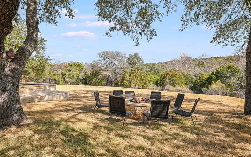 A spacious backyard with a circular firepit surrounded by chairs and outdoor lighting, featuring lush green grass and mature trees.