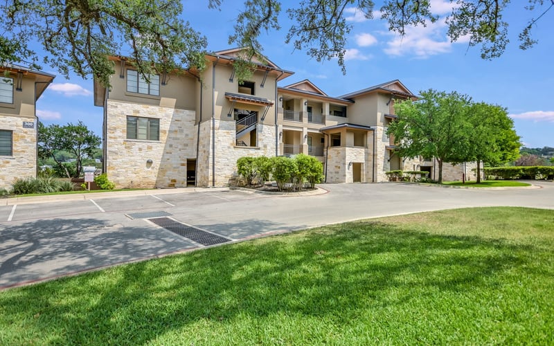 Modern multi-story residential building with stone and stucco exterior, manicured lawn, parking area, and mature trees overhead.