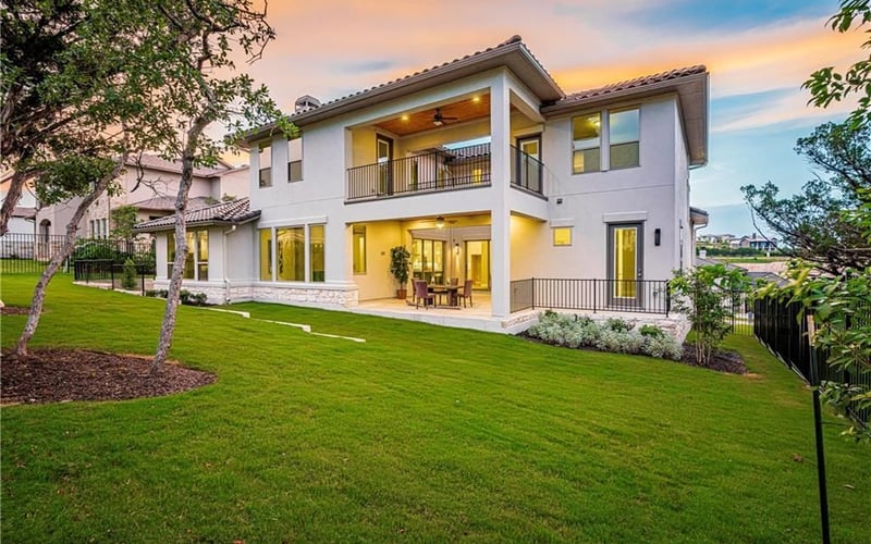 Modern two-story white house with stone base, lit interior, second-floor balcony, and manicured lawn at sunset.